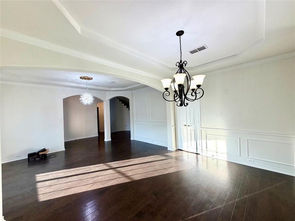 Tray ceilings with crown molding, dark wood-finish flooring, decorative wall paneling, and intricate metal chandeliers