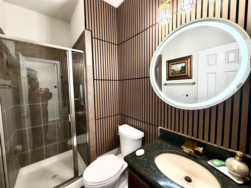 Bathroom featuring vertical wood-slat wall paneling, an oval LED-backlit mirror, dark stone-finish countertop, integrated vanity sink with brass-tone faucet, and a glass-enclosed shower