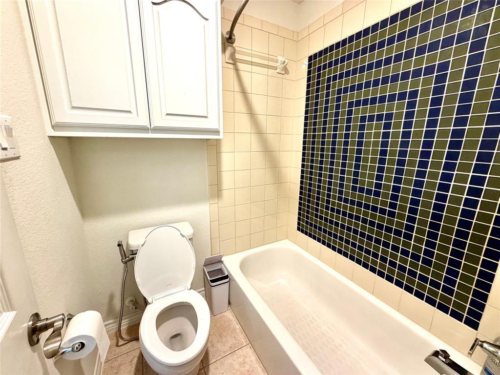 Full bathroom featuring a tub-shower combination with geometric multi-colored wall tile, cream-toned square tile surround, built-in white upper cabinetry, and a high-profile commode with an attached handheld sprayer