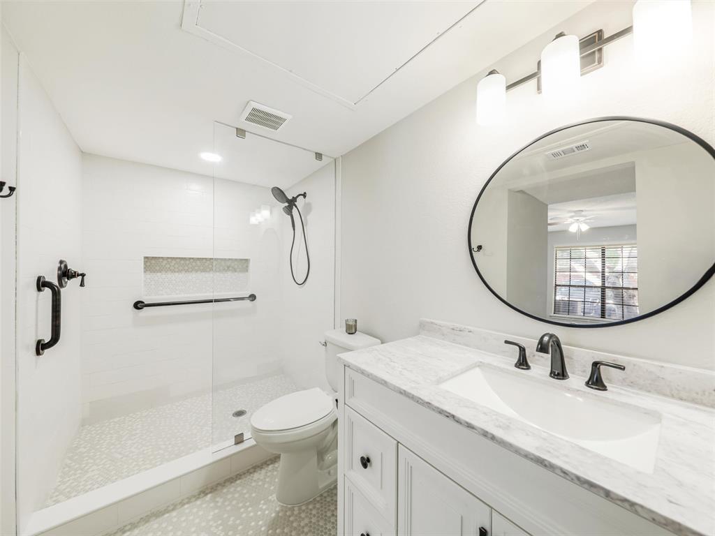 White vanity with a stone countertop, oval mirror, walk-in shower with a glass panel, stone tile flooring, and matte black fixtures