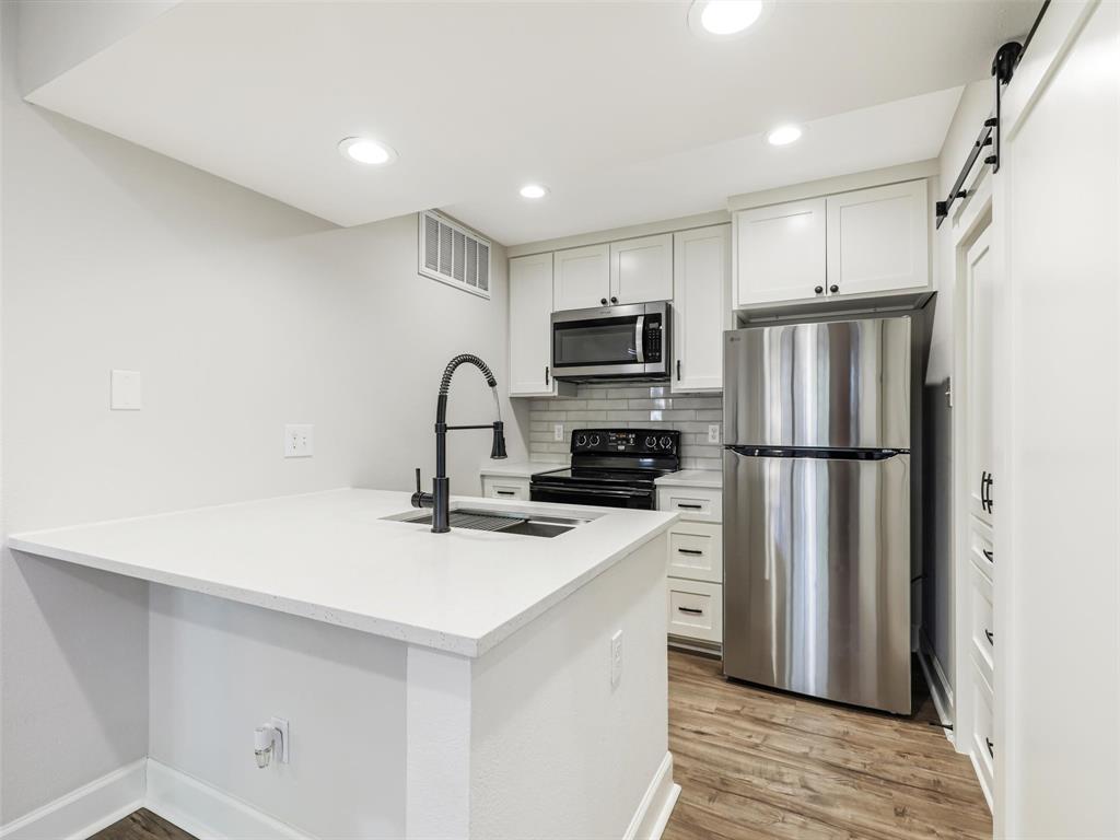 Kitchen featuring white shaker-style cabinetry, solid-surface countertops, stainless steel appliances, matte black plumbing fixtures, and wood-finish flooring Kitchen featuring white shaker-style cabinetry, solid-surface countertops, stainless steel appliances, matte black plumbing fixtures, and wood-finish flooring