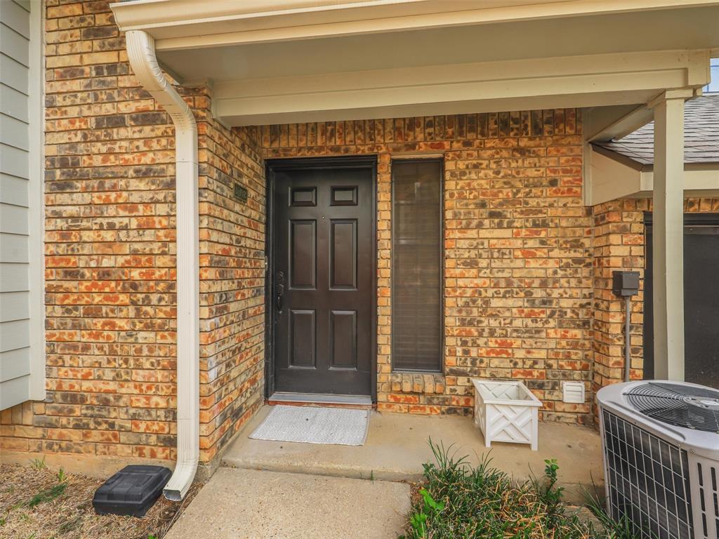 Exterior entryway featuring a multi-tone brick facade, six-panel dark entry door, sidelight window, and a covered porch with concrete flooring Exterior entryway featuring a multi-tone brick facade, six-panel dark entry door, sidelight window, and a covered porch with concrete flooring