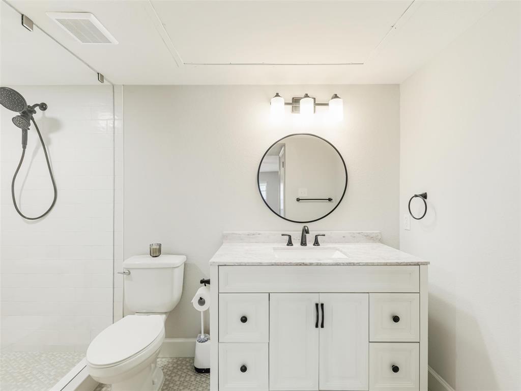 Bathroom featuring a white vanity with stone-top surface, undermount sink, round wall-mounted mirror, three-light fixture, and a glass-enclosed shower with handheld sprayer