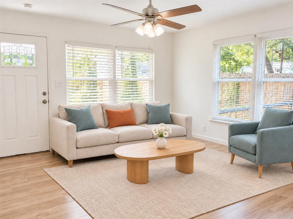Living area featuring wood-finish flooring, white walls, multiple windows with blinds, a ceiling fan with integrated light fixture, and an exterior door with a leaded glass insert