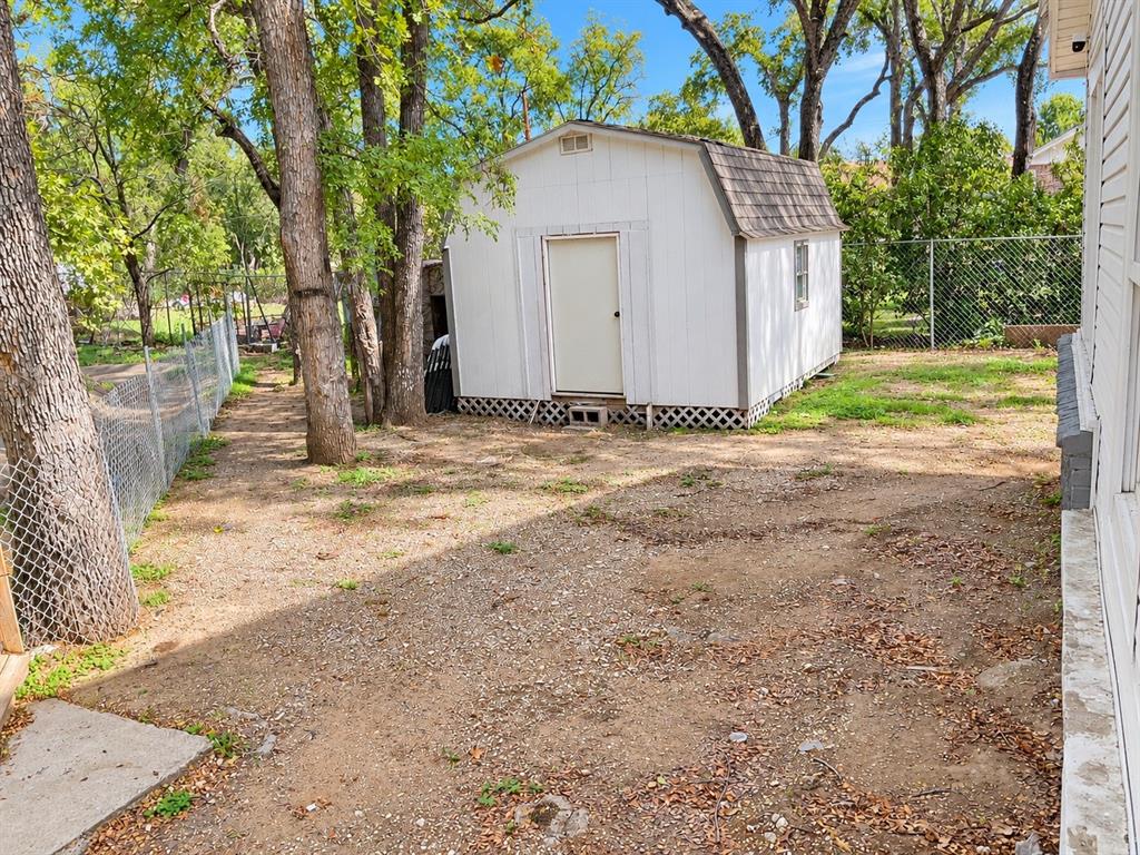 Detached storage shed with a gambrel roof, set within a fenced outdoor space featuring mature trees and lattice underpinning