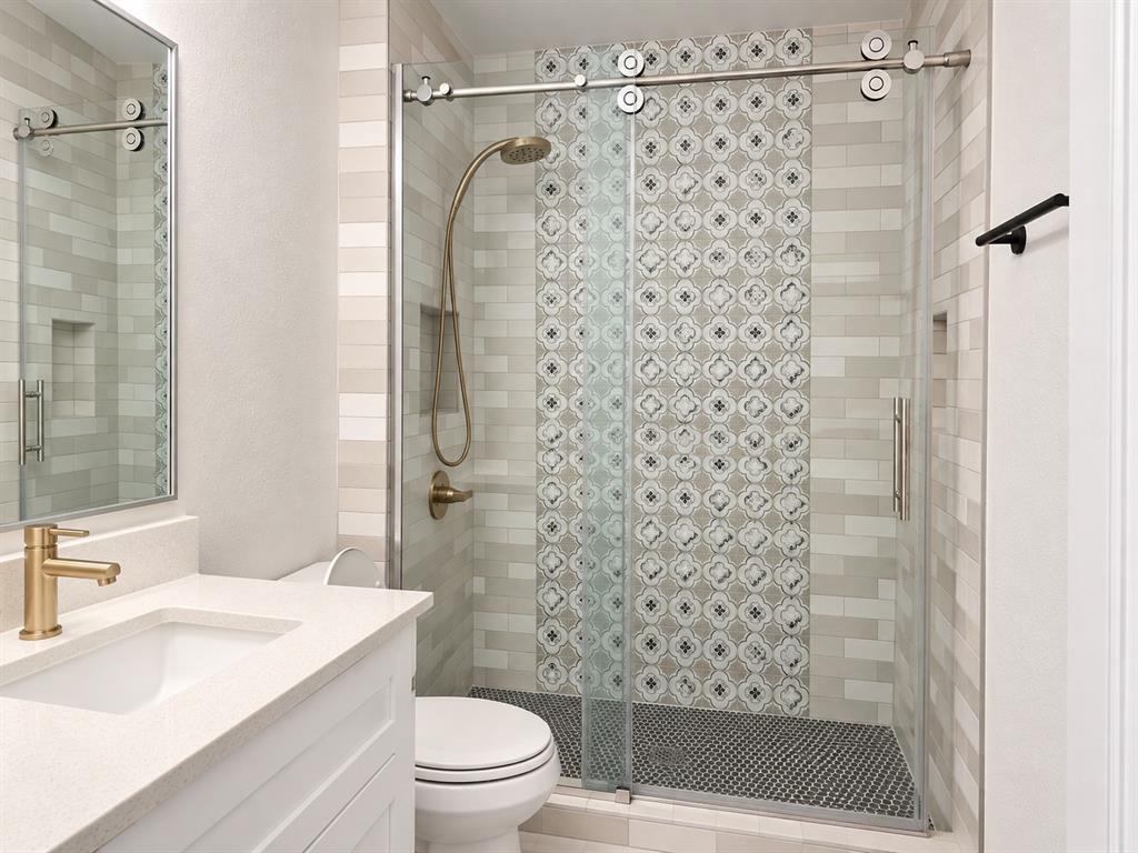 Bathroom featuring a white vanity with a neutral solid-surface countertop, gold-toned faucet, walk-in shower with sliding glass door, decorative patterned tile accent wall, and circular floor tile