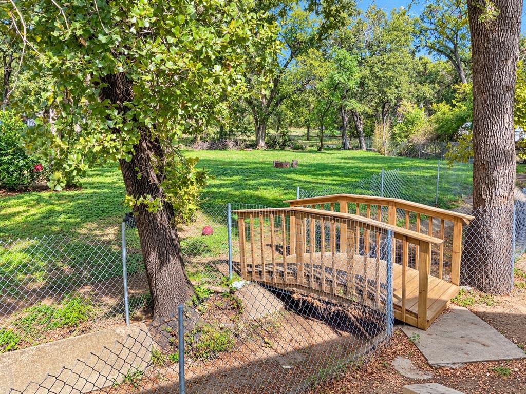 Expansive backyard featuring a manicured lawn, mature trees, chain-link perimeter fencing, and a custom wooden arched footbridge