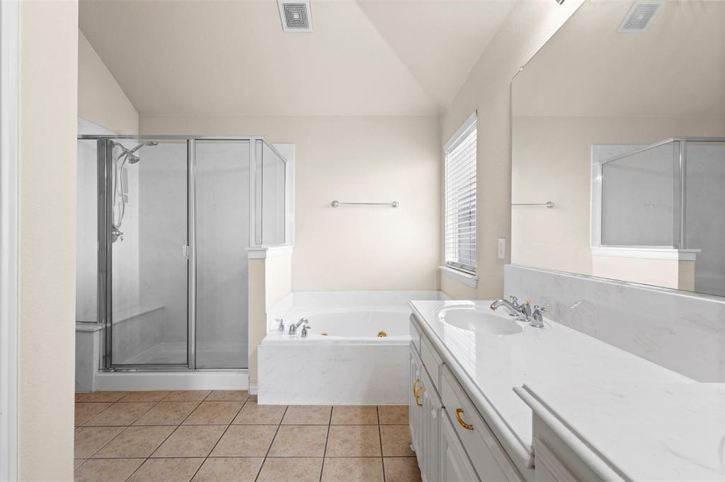 Bathroom featuring a dual-sink vanity with a solid-surface countertop, a drop-in soaking tub with a stone-finish surround, a glass-enclosed shower, and ceramic tile flooring