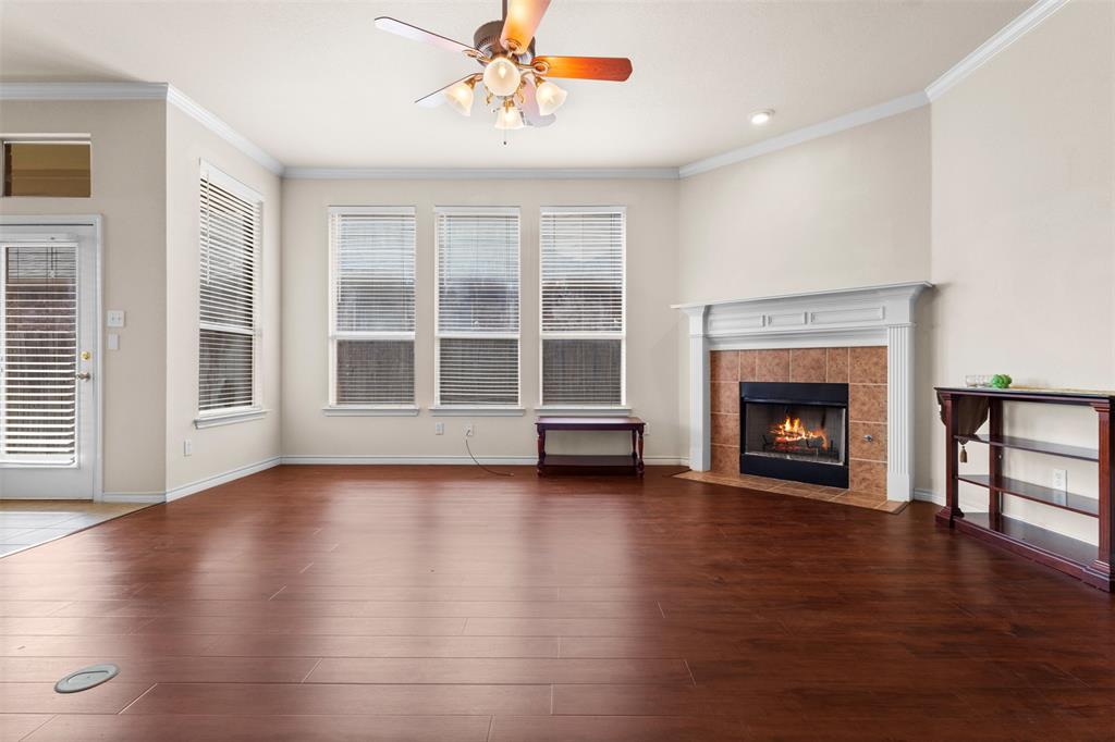 Living area featuring wood-finish flooring, a fireplace with a white mantel and tile surround, crown molding, and multiple windows with horizontal blinds