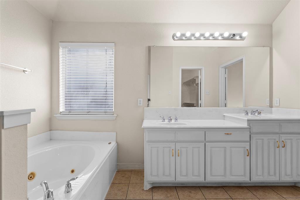 Dual-sink vanity with white cabinetry and stone-finish countertop, jetted soaking tub, oversized wall-mounted mirror, and tile flooring