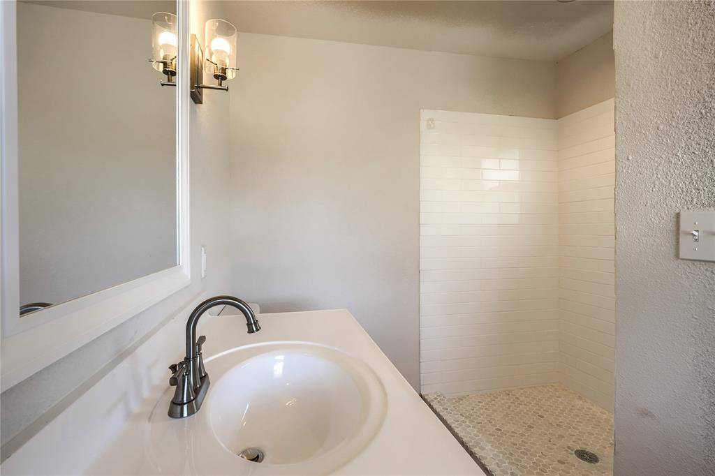 Bathroom vanity featuring a white countertop with an integrated oval sink, metallic gooseneck faucet, wall-mounted light fixture with glass shades, and a shower enclosure with white subway tile surround and mosaic floor tiling