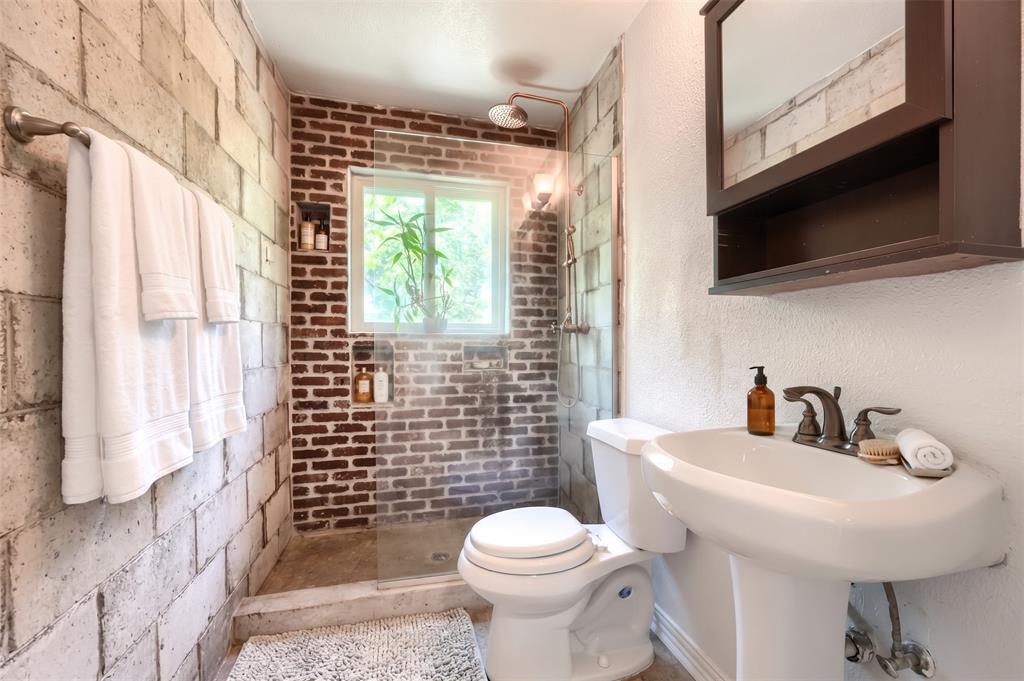 Bathroom featuring exposed cinder block and red brick walls, walk-in shower with glass panel, pedestal sink with multi-handle faucet, and built-in wall-mounted medicine cabinet