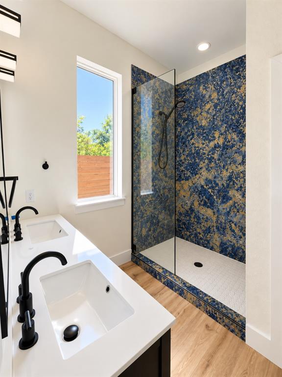 Dual-sink vanity with white countertops and matte black fixtures, walk-in shower featuring blue patterned tile surround and glass enclosure, and wood-finish flooring