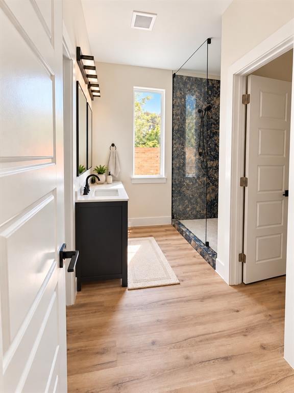 Bathroom featuring wood-finish flooring, a charcoal-toned vanity with a white countertop, a walk-in shower with a glass enclosure and decorative tile surround, and matte black fixtures