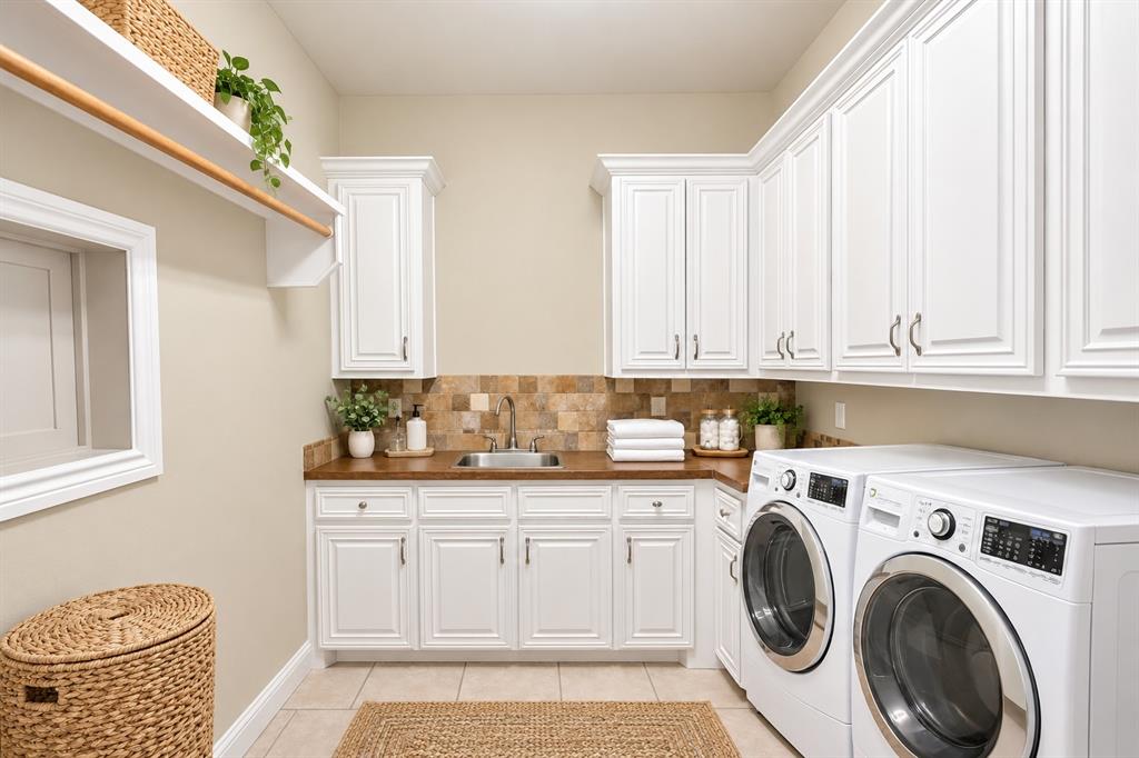 Laundry room featuring white shaker-style cabinetry, wood-finish countertops, tile backsplash, stainless steel drop-in sink, and tile flooring