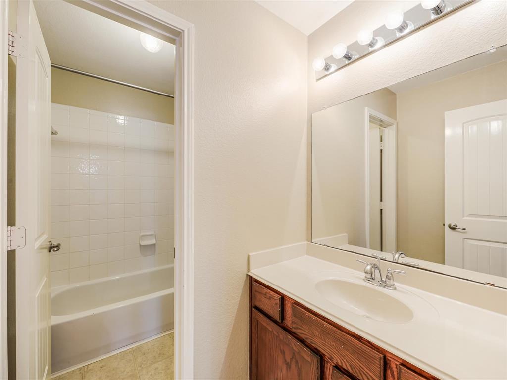 Bathroom featuring a vanity with wood-finish cabinetry, a white integrated countertop and basin, a full-width mirror, and a wall-mounted light fixture, with a separate tub and shower enclosure tiled in white ceramic