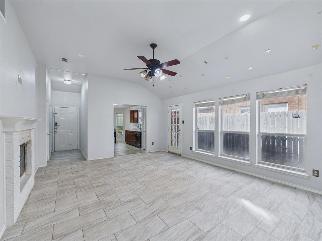 Vaulted ceiling featuring a central light-integrated fan, white-painted brick fireplace, neutral stone-finish tile flooring, and large windows providing natural light