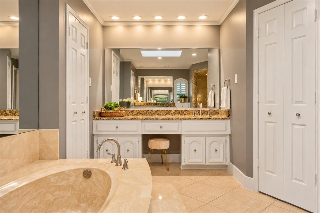 Custom vanity featuring white cabinetry, stone countertop, and integrated makeup station, complemented by a stone-surround soaking tub, large wall-mounted mirror, recessed lighting, and tile flooring