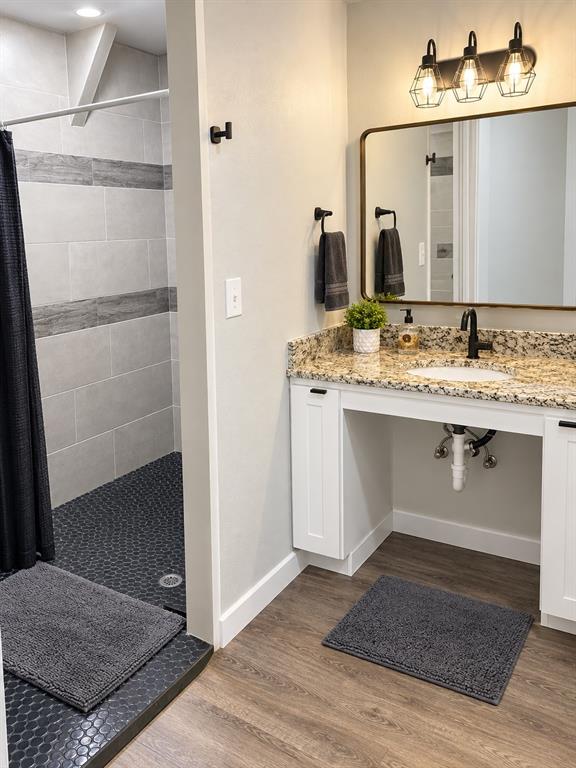 Bathroom featuring a granite countertop, white shaker-style cabinetry, a brass-framed rectangular mirror, three-light industrial vanity fixture, and a tiled shower stall with hex-patterned flooring