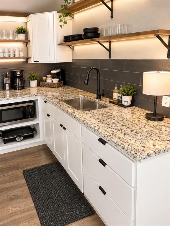 Kitchen featuring white cabinetry with matte black hardware, stone-finish countertops, dark rectangular tile backsplash, wood-finish shelving with under-cabinet lighting, and a stainless steel undermount sink with a matte black faucet