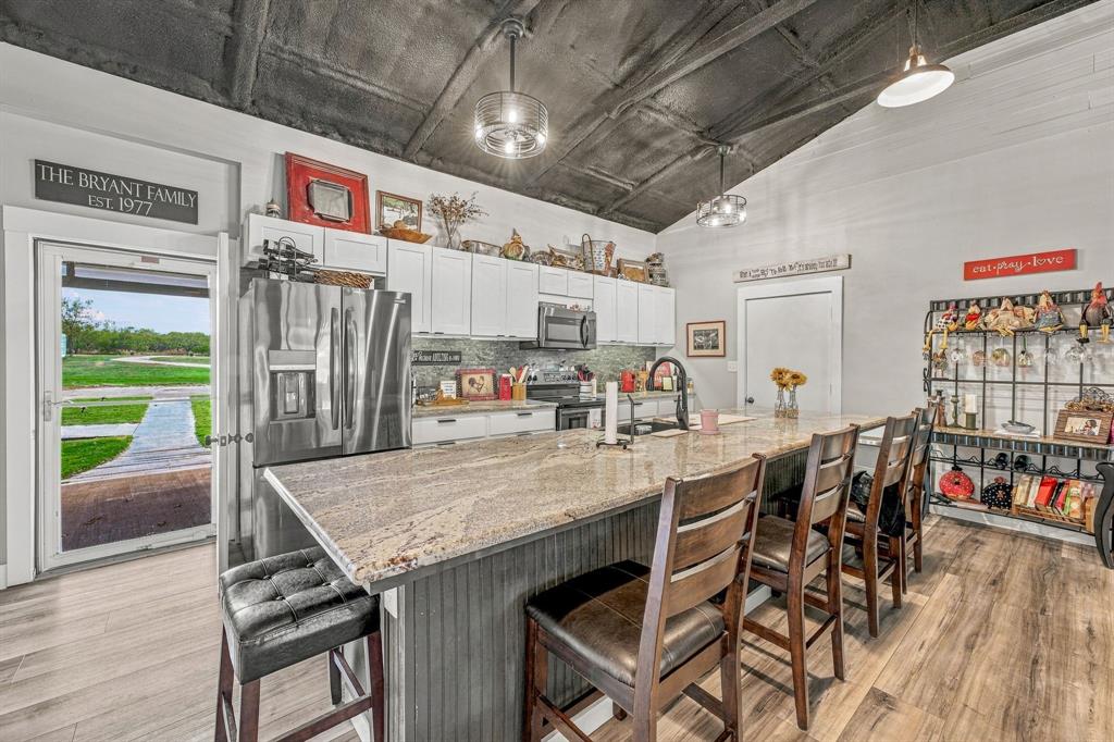 Kitchen featuring wood-finish flooring, granite-topped center island with vertical slat cabinetry, white upper and lower cabinetry, stainless steel appliances, and vaulted ceiling with exposed rafters