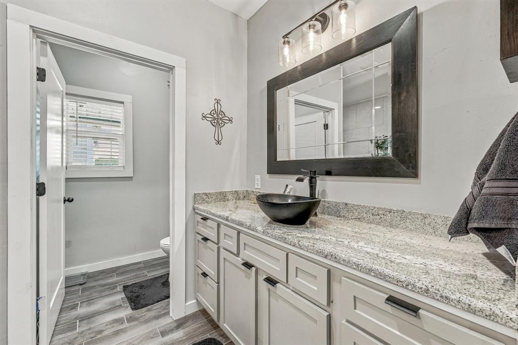 Light-toned vanity cabinetry topped with granite-finish countertop, dark vessel sink, matte black faucet, and wood-finish tile flooring