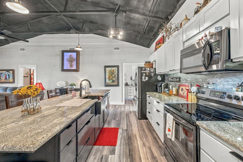 Kitchen featuring granite countertops, an expansive center island with an integrated farmhouse sink, wood-finish flooring, white shaker-style cabinetry, and vaulted ceilings with exposed beams