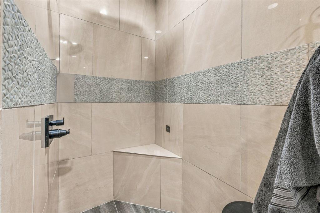 Walk-in shower featuring large-format beige tile, decorative horizontal stone-texture accent inlay, built-in corner bench, and matte black wall-mounted shower fixtures