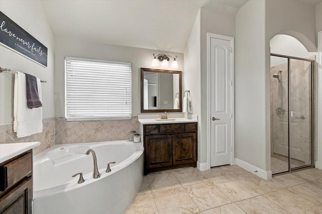 Primary Bathroom features a vaulted ceiling, a vanity light fixture, a corner garden tub with tile surround beneath a window, a linen closet, and a separate walk-in shower with glass enclosure set within an arched opening. Ceramic tile flooring extends throughout.