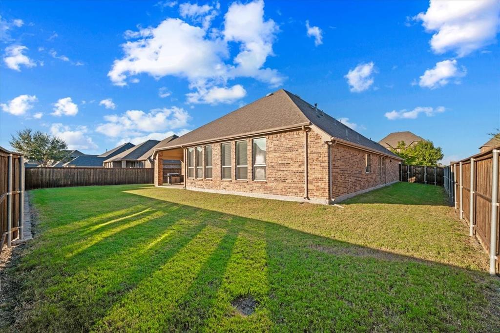 Backyard features a view of the hip roofline and brick construction with gutters, a row of windows along the back of the home, wood privacy fencing with metal posts, and an open grass lawn.