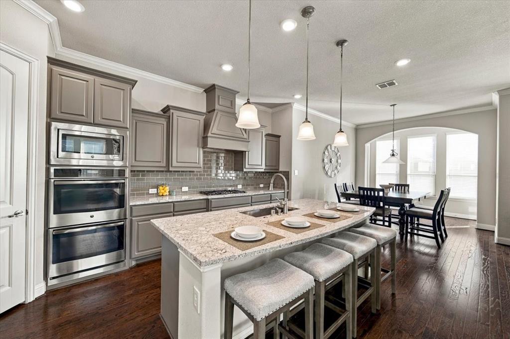 Kitchen features gray cabinetry, a built-in stainless microwave and double wall oven, countertops along the perimeter and island, and an open layout to the dining area. Wood flooring extends throughout.