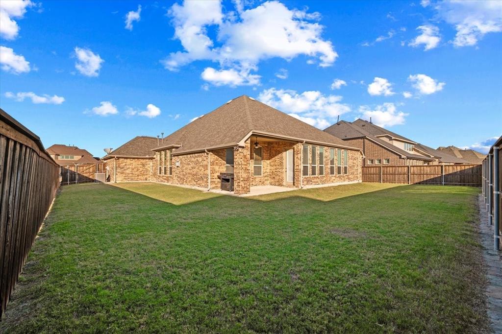 Backyard features a view of the composition roof and brick construction, a covered patio, multiple windows, wood privacy fencing with metal posts, an underground sprinkler system, and a spacious grass lawn.