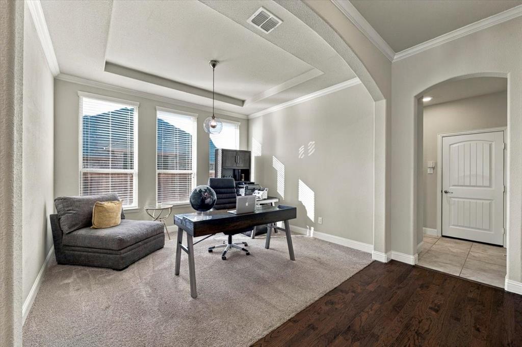 Office features a tray ceiling with crown molding, a pendant light fixture, three windows, and arched openings to the main hall and the adjacent hall. Carpet flooring extends throughout, transitioning to wood and ceramic tile in the adjoining halls. Office features a tray ceiling with crown molding, a pendant light fixture, three windows, and arched openings to the main hall and the adjacent hall. Carpet flooring extends throughout, transitioning to wood and ceramic tile in the adjoining halls.