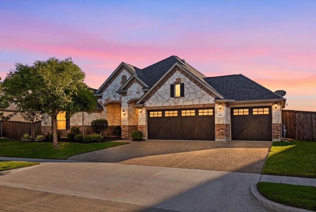 Front exterior features a composition roof with gabled rooflines, stone and brick construction, coach lights, an arched window with shutters, an attached three-car garage with carriage-style doors, a mature tree, landscaped beds, and a concrete driveway. Front exterior features a composition roof with gabled rooflines, stone and brick construction, coach lights, an arched window with shutters, an attached three-car garage with carriage-style doors, a mature tree, landscaped beds, and a concrete driveway.