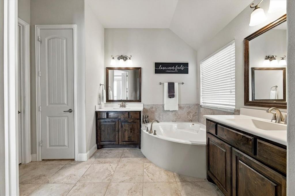 Primary Bathroom features a vaulted ceiling, vanity light fixtures, separate vanities with dark wood cabinetry, a corner garden tub with tile surround beneath a window, and a linen closet. Ceramic tile flooring extends throughout.