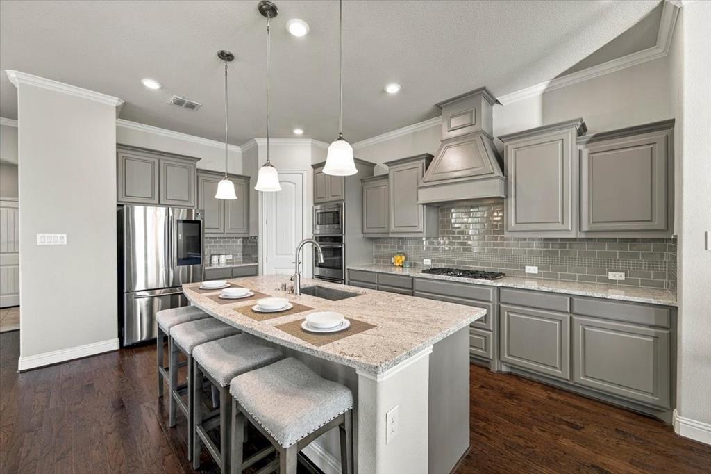 Kitchen features crown molding, three pendant lights over the island, a decorative vent hood, upper cabinets with crown trim, and a curved island with breakfast bar seating. Wood flooring extends throughout.