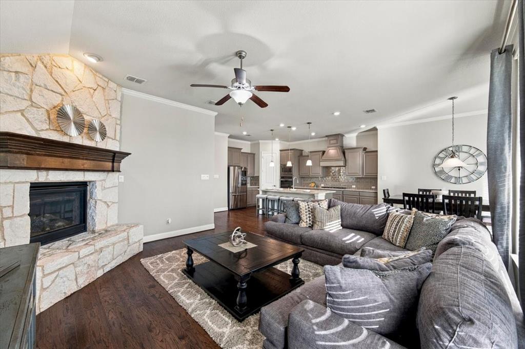 Living Room features crown molding, a ceiling fan with light fixture, a corner gas log fireplace with gas starter, a stone surround and wood mantel, and an open layout connecting to the kitchen and dining area. Wood flooring extends throughout. Living Room features crown molding, a ceiling fan with light fixture, a corner gas log fireplace with gas starter, a stone surround and wood mantel, and an open layout connecting to the kitchen and dining area. Wood flooring extends throughout.