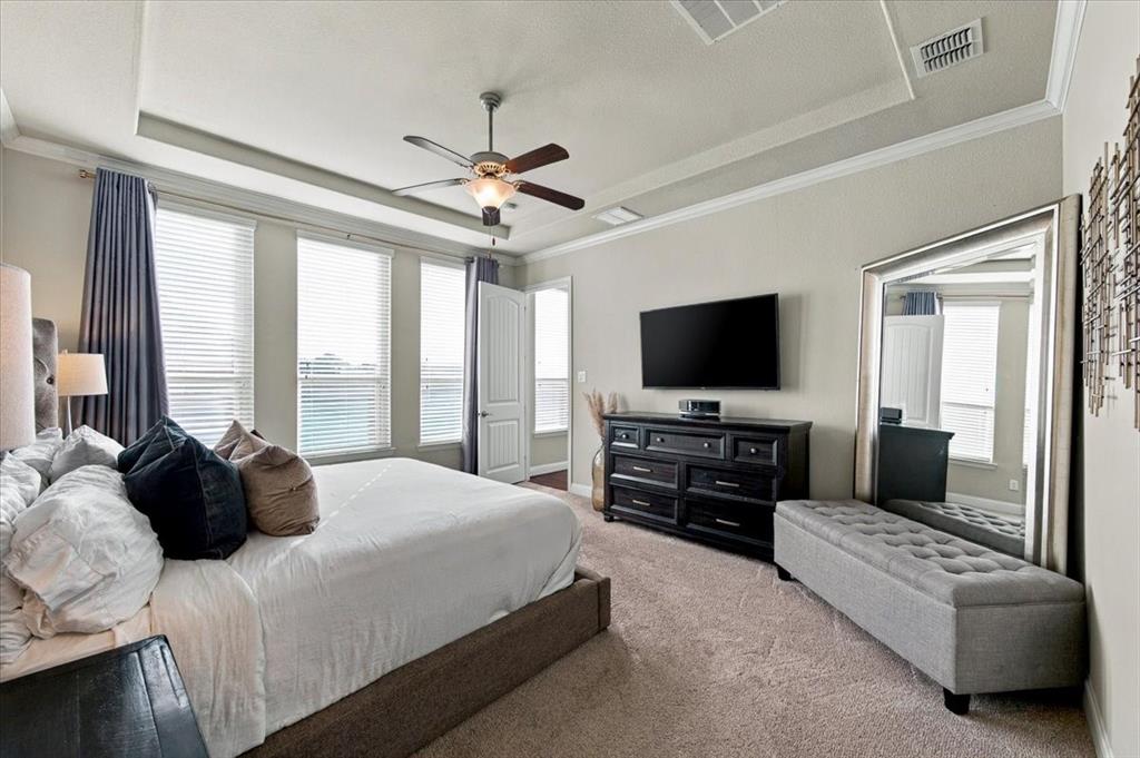 Primary Bedroom features a tray ceiling with crown molding, a ceiling fan with light fixture, a row of three windows, and an entry door. Carpet flooring extends throughout.