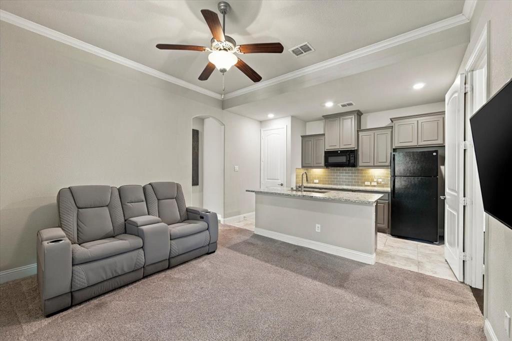 Suite Sitting Room features crown molding and a ceiling fan with light fixture, an arched opening to the suite hall, and an open layout to the kitchenette. Carpet flooring extends throughout, transitioning to ceramic tile in the kitchenette.