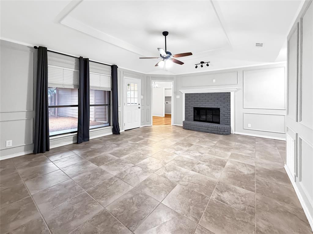 Living area featuring large-format tile flooring, a painted brick fireplace with mantel, recessed tray ceiling, decorative wall molding, and a central ceiling fan