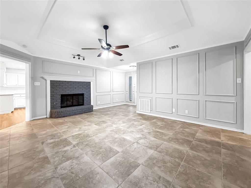 Tray ceiling featuring a central ceiling fan, brick fireplace with a decorative mantel, and wall wainscoting complemented by large-format tile flooring
