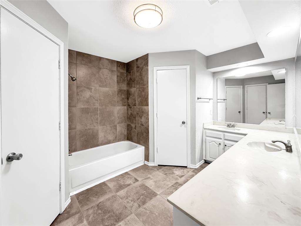 Bathroom featuring dual-sink vanity with light-toned solid surface countertop, tub with full-height tile surround, and tile flooring