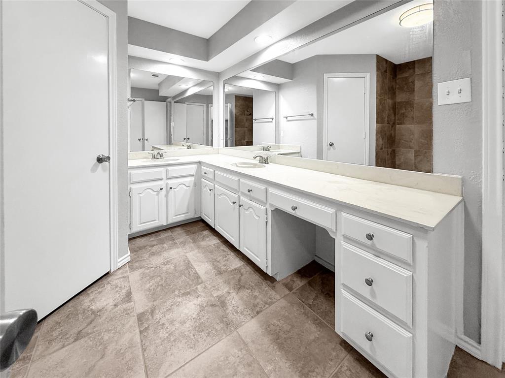 L-shaped vanity featuring white cabinetry, a solid surface countertop, dual integrated sinks with chrome fixtures, and a full-width vanity mirror set against tile flooring