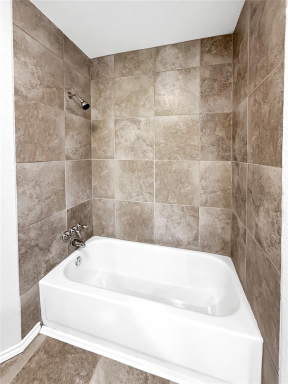 Bathtub surround featuring floor-to-ceiling square earth-tone wall tiles, a built-in white acrylic tub, and polished chrome plumbing fixtures