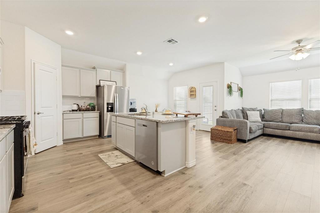 Open-concept kitchen featuring shaker-style cabinetry, stone countertops, a central island with an undermount sink, stainless steel appliances, and wood-finish flooring throughout
