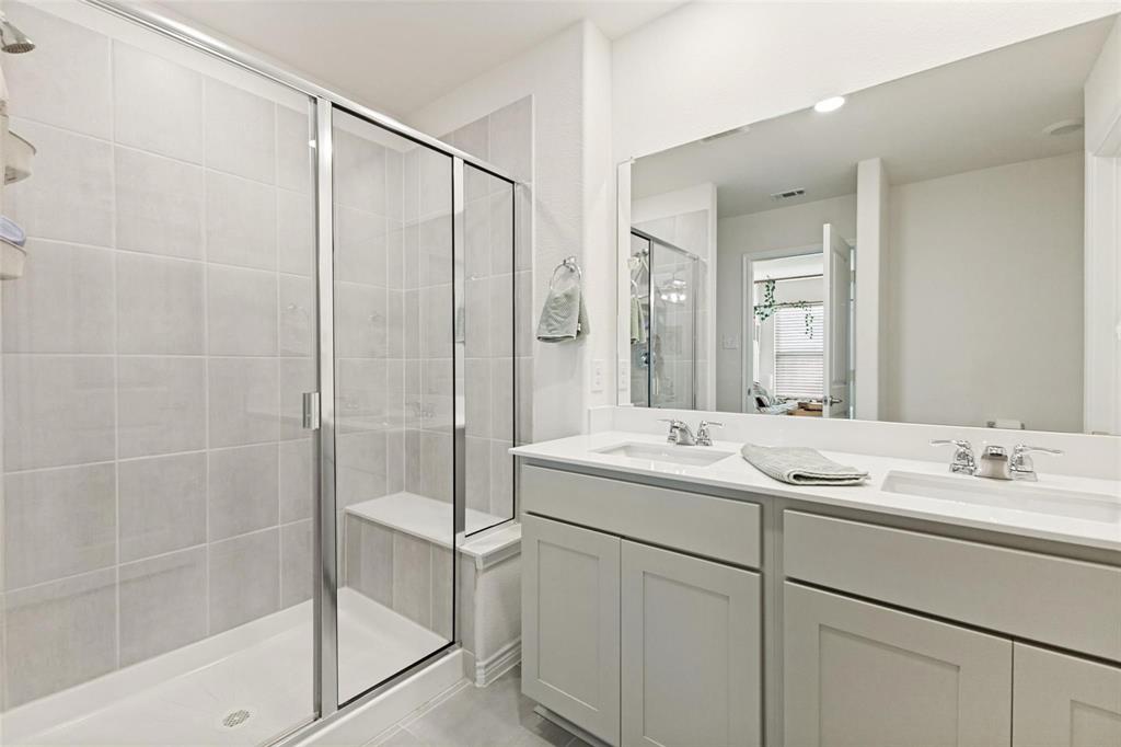 Bathroom featuring a dual-sink vanity with solid-surface countertop, full-width mirror, and a walk-in shower with tiled surround and integrated bench