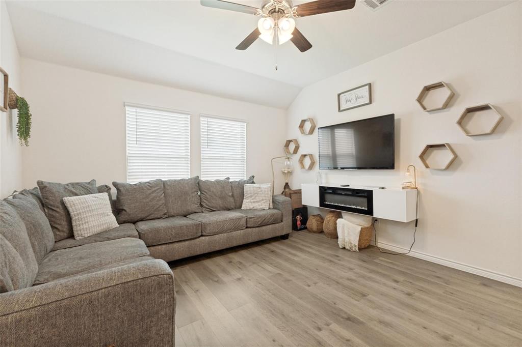 Living area featuring wood-finish flooring, vaulted ceiling with a central paddle fan, dual windows with horizontal blinds, and a wall-mounted floating cabinet with an integrated electric fireplace