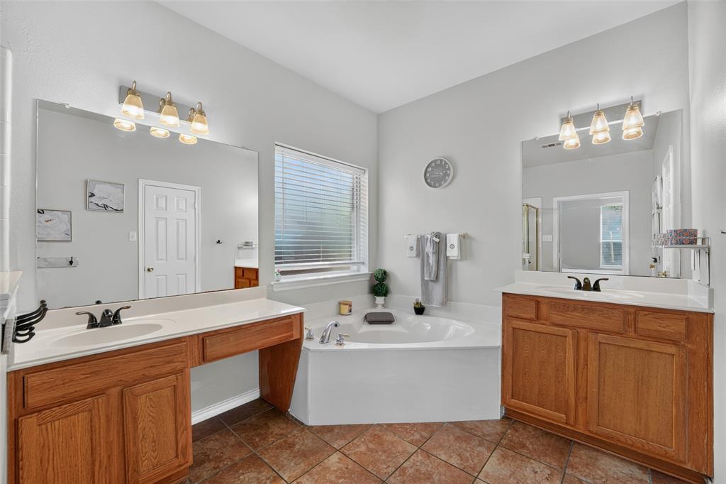 Dual wood-finish vanities with light-toned countertops, corner soaking tub, ceramic tile flooring, and large wall-mounted mirrors