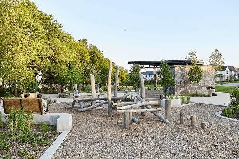 Outdoor recreational area featuring wood-beam climbing structures, integrated concrete bench seating, wood chip surfacing, and a stone-clad pavilion
