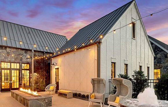 Modern farmhouse exterior featuring vertical white board-and-batten siding, stone masonry accents, and a dark metal roof, complemented by a paver patio with a built-in linear stone fire feature